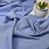 Un tissu seersucker uni texturé au mètre de Longan Craft France, doux et violet clair, est drapé à côté d'un petit pot en céramique blanc contenant une succulente verte et des galets blancs.