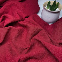 Un tissu polyester seersucker rouge texturé de Longan Craft France est présenté avec des plis, tandis qu'un petit pot en céramique beige avec une succulente verte et des pierres blanches se trouve dans le coin supérieur droit.