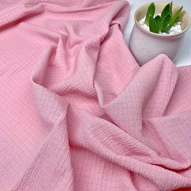 Le Tissu seersucker uni texturé au mètre de Longan Craft France, présenté en plis doux, est exposé à côté d'une petite plante verte dans un pot en céramique légère avec des pierres blanches.