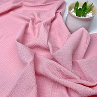 Le Tissu seersucker uni texturé au mètre de Longan Craft France, présenté en plis doux, est exposé à côté d'une petite plante verte dans un pot en céramique légère avec des pierres blanches.