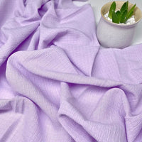 Un tissu seersucker Vichy violet clair de Longan Craft France aux plis délicats est posé à côté d'une petite plante succulente en pot dans un pot en céramique blanc rempli de pierres. Tissu seersucker uni texturé, froissé polyester, à carreaux au mètre.