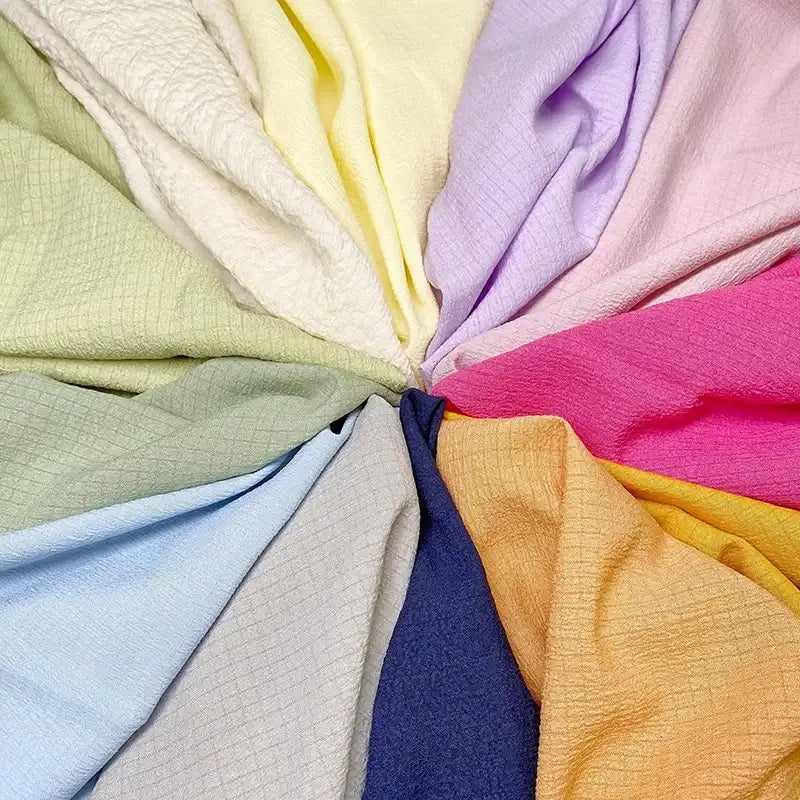 Les tissus seersucker de Longan Craft France -uni, polyester froissé et motifs à carreaux- sont présentés dans un cercle aux couleurs distinctes : crème, jaune, violet clair, rose, rouge, orange, bleu marine, gris, bleu clair et vert.