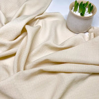Un tissu seersucker texturé à carreaux de couleur crème de Longan Craft France est étalé, avec une petite plante en pot dans des galets blancs placée en haut à droite. Tissu seersucker uni texturé au mètre | Polyester | à carreaux.