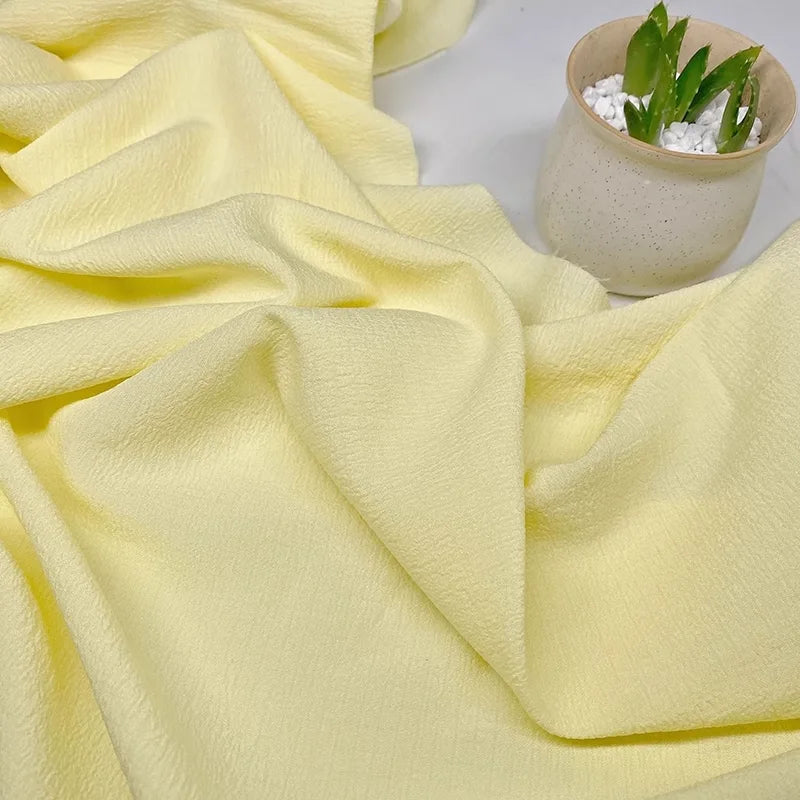 Un tissu texturé en polyester à carreaux seersucker jaune clair de Longan Craft France est présenté à côté d'un pot beige avec une succulente verte et des galets blancs.