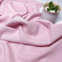 Un morceau de tissu en polyester seersucker rose pâle de Longan Craft France, à la finition texturée et froissée, est étalé à côté d'un petit pot blanc contenant une succulente verte et des galets blancs.