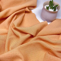 Le Tissu seersucker à carreaux orange de Longan Craft France, légèrement froissé, est étalé à côté d'une petite plante en pot céramique clair rempli de galets blancs.