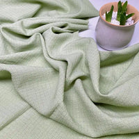 Le Tissu seersucker uni texturé de Longan Craft France, en polyester vichy vert clair, est doucement drapé sur une surface où un pot en céramique contenant des pierres blanches et des plantes grasses est placé en haut à droite.