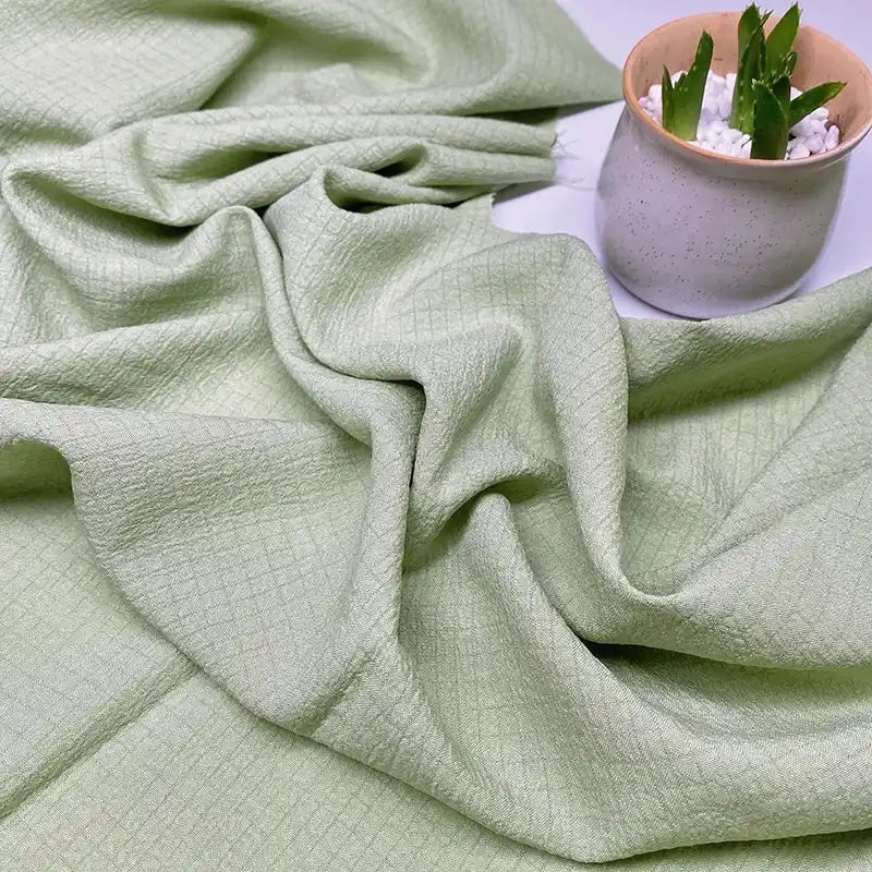 Le Tissu seersucker uni texturé de Longan Craft France, en polyester vichy vert clair, est doucement drapé sur une surface où un pot en céramique contenant des pierres blanches et des plantes grasses est placé en haut à droite.