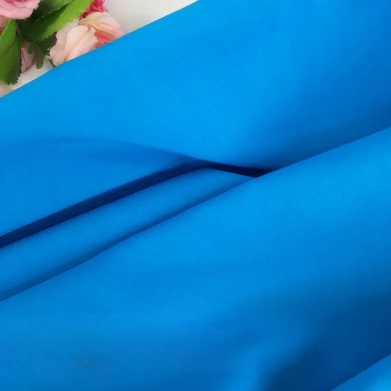 Gros plan sur la Mousseline extensible au mètre de Longan Craft France, bleu vif et lisse, avec des plis doux ; des fleurs artificielles roses partiellement visibles avec des feuilles vertes apparaissent dans le coin supérieur gauche.