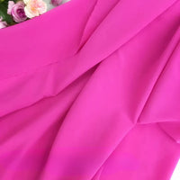 Un gros plan de Longan Craft France Mousseline polyester épaisse et extensible montre un tissu rose vif avec des plis doux et une texture visible. Des fleurs artificielles rose pâle et blanches sont partiellement visibles dans le coin supérieur gauche.