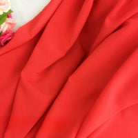 Un gros plan de la Mousseline rouge vif extensible au mètre de Longan Craft France montre des plis doux, avec une grappe de fleurs artificielles roses et blanches dans le coin.