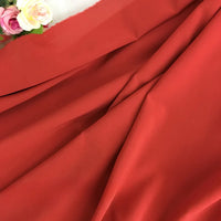 Un gros plan de la Mousseline rouge extensible au mètre de Longan Craft France montre ses plis lisses et légèrement froncés, avec des fleurs artificielles roses et blanches dans le coin supérieur gauche. Il s'agit d'une mousseline de polyester épaisse et extensible.