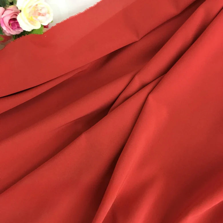 Un gros plan de la Mousseline rouge extensible au mètre de Longan Craft France montre ses plis lisses et légèrement froncés, avec des fleurs artificielles roses et blanches dans le coin supérieur gauche. Il s'agit d'une mousseline de polyester épaisse et extensible.