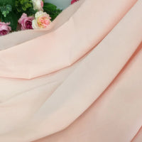 Un morceau de Mousseline rose pâle extensible au mètre de Longan Craft France est drapé de plis, avec des fleurs artificielles roses et vertes partiellement visibles à l'arrière-plan dans le coin supérieur gauche.
