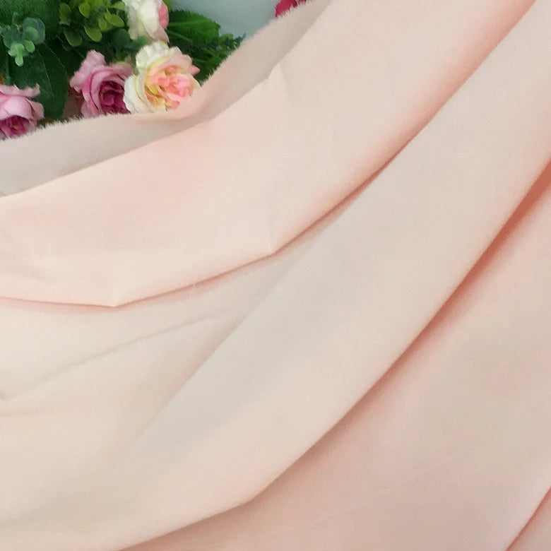 Un morceau de Mousseline rose pâle extensible au mètre de Longan Craft France est drapé de plis, avec des fleurs artificielles roses et vertes partiellement visibles à l'arrière-plan dans le coin supérieur gauche.
