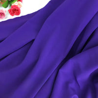 Un gros plan montre Mousseline extensible au mètre de Longan Craft France en polyester violet drapé avec une texture lisse, avec des roses artificielles rouges et roses reposant sur une surface blanche dans le coin supérieur gauche.