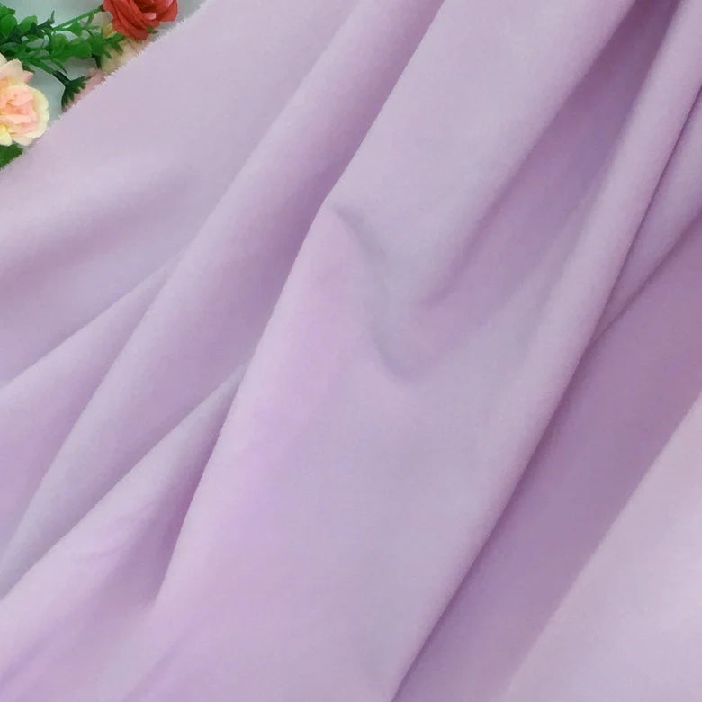 Une douce Mousseline lavande extensible au mètre de Longan Craft France est drapée en diagonale sur l'image, avec quelques fleurs artificielles roses, jaunes et orange partiellement visibles dans le coin supérieur gauche.