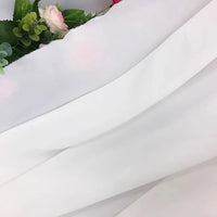 Gros plan de la Mousseline extensible au mètre (Mousseline polyester épaisse et extensible) de Longan Craft France, drapée en diagonale et recouvrant partiellement des fleurs artificielles et des feuilles vertes dans le coin supérieur gauche.