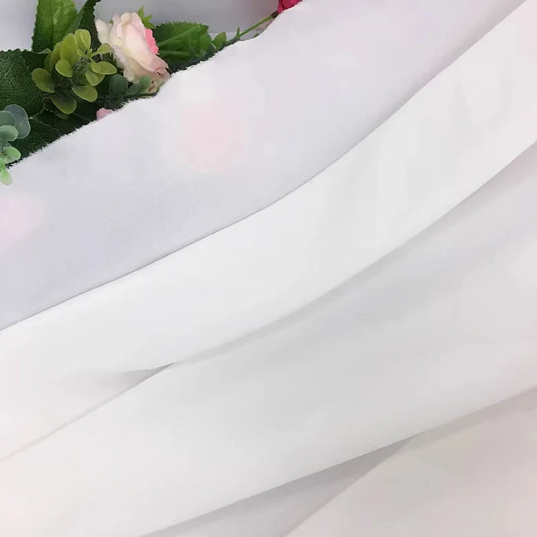 Gros plan de la Mousseline extensible au mètre (Mousseline polyester épaisse et extensible) de Longan Craft France, drapée en diagonale et recouvrant partiellement des fleurs artificielles et des feuilles vertes dans le coin supérieur gauche.