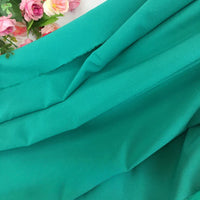 Gros plan sur la Mousseline extensible au mètre-épaisseur et extensible polyester de Longan Craft France, avec un tissu turquoise brillant plié en douceur et un groupe de fleurs artificielles roses dans le coin supérieur gauche. Finition mate.