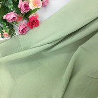 Un morceau de Mousseline vert clair extensible au mètre de Longan Craft France, aux plis apparents, est déployé ; des roses artificielles roses et violettes reposent dans le coin supérieur gauche.