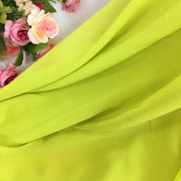 Gros plan sur la Mousseline extensible au mètre de Longan Craft France, de couleur vert lime, drapée de plis souples, avec des fleurs artificielles roses et blanches disposées dans le coin supérieur gauche.
