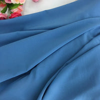 Gros plan sur la Mousseline polyester épaisse et extensible au mètre de Longan Craft France en bleu lisse, posée à plat, avec des fleurs artificielles roses et rouges partiellement visibles dans le coin supérieur gauche.