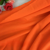 La Mousseline orange vif extensible au mètre de Longan Craft France est élégamment drapée, avec des roses artificielles roses et rouges en haut à gauche. C'est le tissu polyester idéal pour les robes de chambre et les projets créatifs.