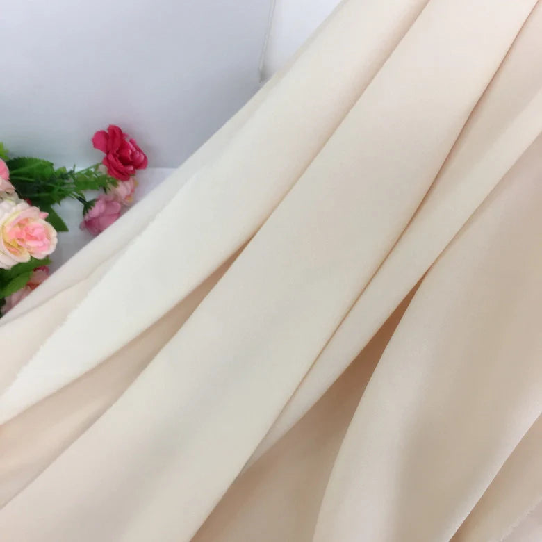 Gros plan sur la Mousseline extensible au mètre de couleur crème de Longan Craft France, un épais tissu de polyester extensible drapé en plis souples avec des fleurs artificielles roses et rouges sur le côté.
