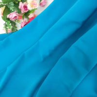 Gros plan sur la Mousseline extensible au mètre de Longan Craft France, en polyester bleu vif à peine froissé. En haut à gauche, on aperçoit un bouquet de fausses roses rose et crème avec feuilles vertes.