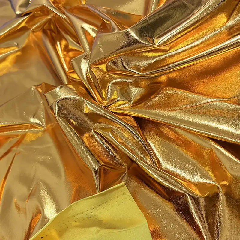 Un gros plan de Tissu lamé stretch au mètre de Longan Craft France montre du polyester doré froissé et brillant avec une finition réfléchissante et des plis, souvent utilisé pour les costumes ou l'artisanat. Un matériau jaune mat est visible en bas.