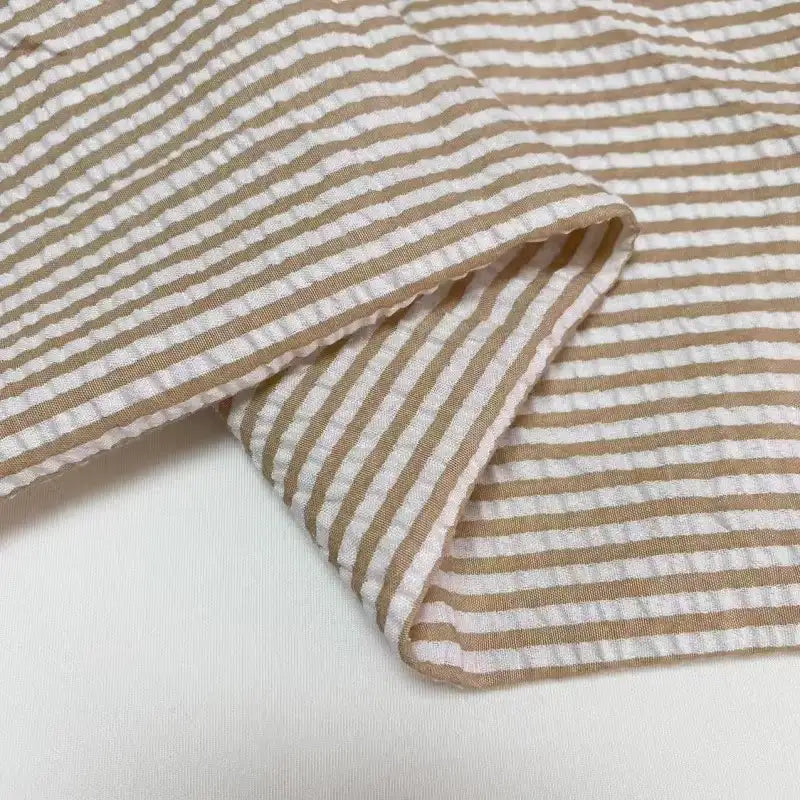 Un gros plan du Seersucker rayé au mètre de Longan Craft France montre son tissu rayé beige et blanc en polyester-coton, plié avec des lignes horizontales - un tissu seersucker idéal pour l'été, parfait pour les vêtements d'été légers et aérés.