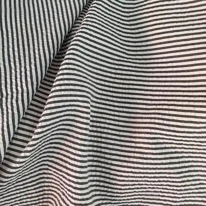 Un gros plan du Seersucker rayé au mètre de Longan Craft France montre ses rayures noires et blanches ondulées, mettant en valeur les plis uniques et la texture légère du polyester-coton - parfait comme tissu seersucker pour l'été.