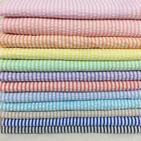Une pile bien empilée de Seersucker rayé au mètre de Longan Craft France présente des rayures horizontales pastel - rose, pêche, jaune, vert, bleu, violet, blanc - qui mettent en valeur ce tissu d'été léger en polyester-coton.