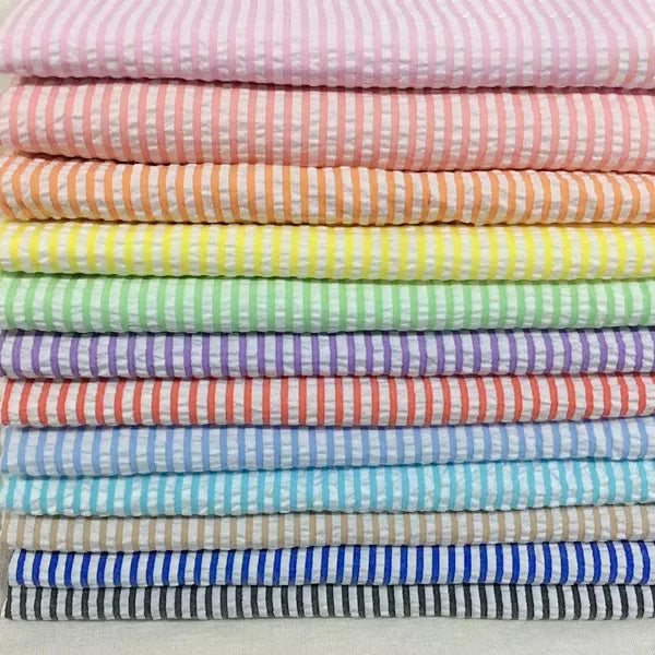Une pile bien empilée de Seersucker rayé au mètre de Longan Craft France présente des rayures horizontales pastel - rose, pêche, jaune, vert, bleu, violet, blanc - qui mettent en valeur ce tissu d'été léger en polyester-coton.