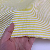 A hand holds Seersucker rayé au mètre de Longan Craft France, un tissu polyester-coton à rayures jaunes et blanches, idéal pour coudre des vêtements d'été légers et confortables.