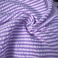 Un gros plan du Seersucker rayé au mètre de Longan Craft France montre son tissu polyester-coton avec des rayures colorées et la texture classique du seersucker - légèrement froissé, froncé, avec une alternance de bandes horizontales violettes et blanches.