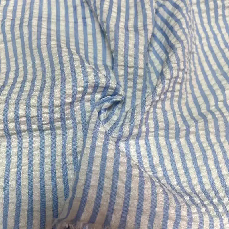Gros plan sur le Seersucker rayé au mètre de Longan Craft France : tissu rayé bleu clair et blanc en polyester-coton, texture douce avec des plis, idéal pour l'été, avec des rayures colorées régulièrement espacées.