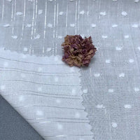 Le Voile plumetis au mètre de Longan Craft France, un tissu transparent en coton blanc à pois suisses avec des rayures argentées, est présenté avec un coin délicatement plié et une fleur rose séchée - idéal pour une décoration intérieure élégante.