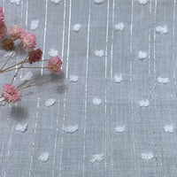 Un gros plan du Voile plumetis au mètre de Longan Craft France montre un tissu de coton blanc avec des fils argentés verticaux et des points en relief, disponible en gris clair ou en blanc. Des fleurs roses séchées ajoutent une touche décorative à la scène.