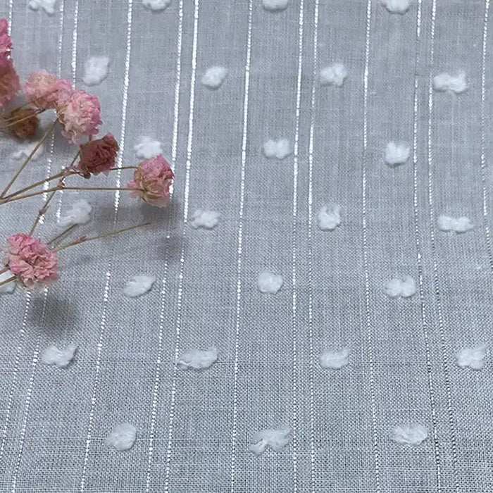 Un gros plan du Voile plumetis au mètre de Longan Craft France montre un tissu de coton blanc avec des fils argentés verticaux et des points en relief, disponible en gris clair ou en blanc. Des fleurs roses séchées ajoutent une touche décorative à la scène.