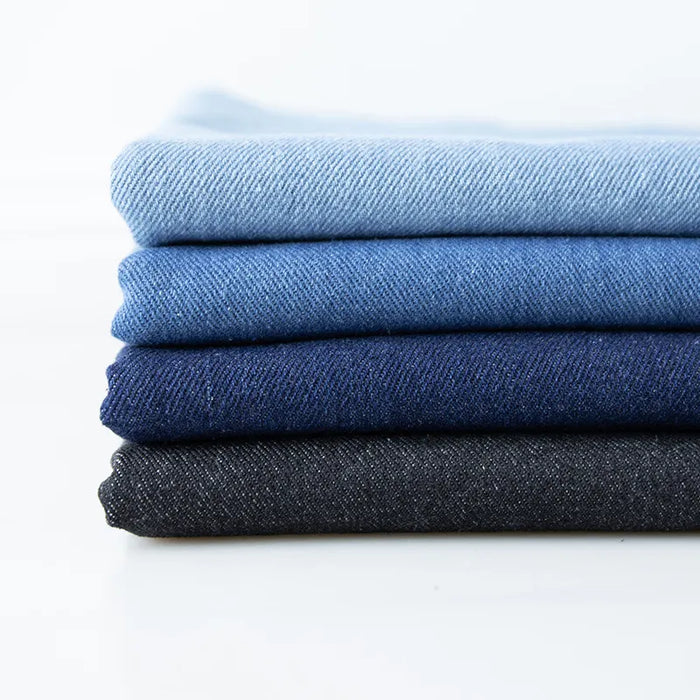 Une pile de quatre tissus en jean coton lavé 14,5 oz de Longan Craft France, dans des tons de denim allant du bleu clair au bleu foncé, est disposée sur une surface blanche unie.