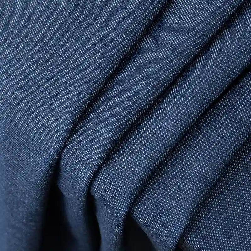 Gros plan sur le Tissu en jean coton lavé 14,5 oz de Longan Craft France : denim bleu foncé de première qualité avec un tissage finement texturé et des mouchetures blanches, présenté en couches avec des plis parallèles. Idéal pour les jeans et disponible au mètre.