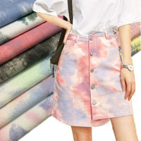 Une personne porte un t-shirt blanc rentré dans une jupe tie-dye en denim pastel fabriquée à partir du "Tissu denim en coton tie-dye au mètre" de Longan Craft France. Des tissus empilés dans différents motifs pastel rehaussent la décoration intérieure.
