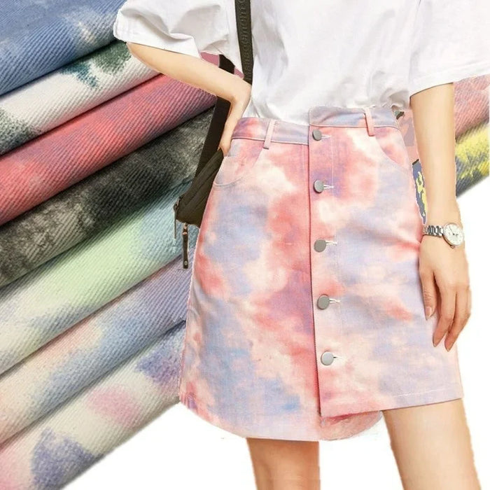Une personne porte un t-shirt blanc rentré dans une jupe tie-dye en denim pastel fabriquée à partir du "Tissu denim en coton tie-dye au mètre" de Longan Craft France. Des tissus empilés dans différents motifs pastel rehaussent la décoration intérieure.