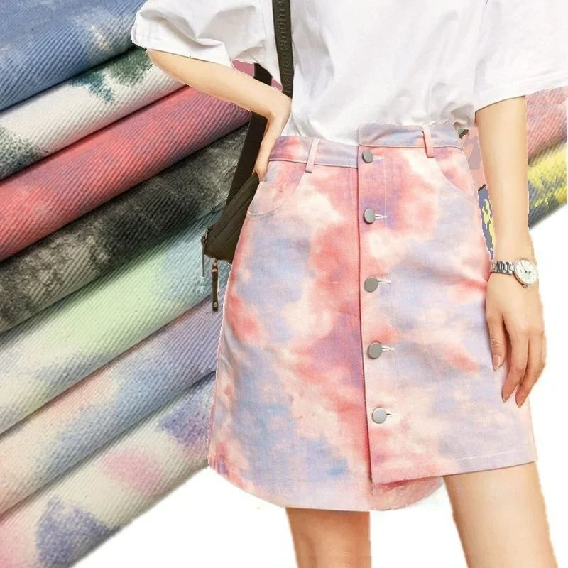 Une personne porte un t-shirt blanc rentré dans une jupe tie-dye en denim pastel fabriquée à partir du "Tissu denim en coton tie-dye au mètre" de Longan Craft France. Des tissus empilés dans différents motifs pastel rehaussent la décoration intérieure.