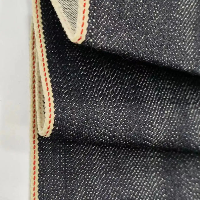 Gros plan sur le Tissu en jean selvedge 16,5 oz au mètre (100 % coton) de Longan Craft France, un denim bleu foncé épais avec une trame blanche visible, des bords de selvage de couleur crème et une ligne de piqûre rouge, soulignant sa texture haut de gamme.