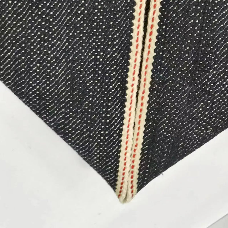 Gros plan sur le Tissu en jean selvedge 16,5 oz de Longan Craft France, un denim 100 % coton épais bleu foncé avec une lisière blanche et rouge, plié pour mettre en valeur la lisière diagonale sur un fond blanc.
