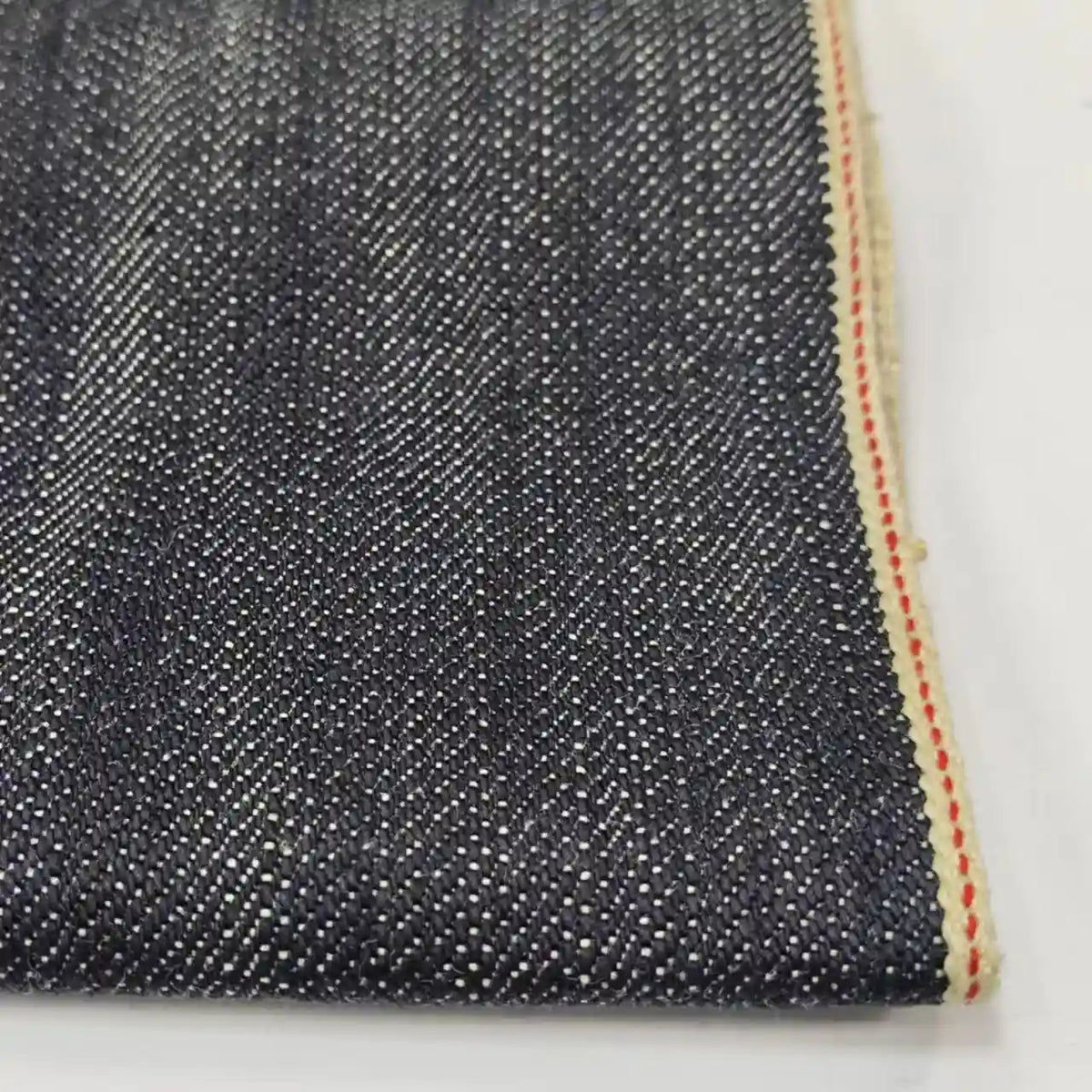 Un gros plan du tissu en jean selvedge 16,5 oz de Longan Craft France (100 % coton) révèle un sergé bleu foncé, des fils de trame blancs et un bord fini avec une bande crème et une ligne rouge. Disponible au mètre comme "Tissu en jean selvedge 16,5 oz.