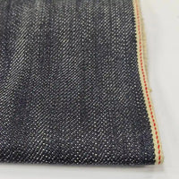 Un gros plan du tissu en jean selvedge 16,5 oz de Longan Craft France (100 % coton) révèle un sergé bleu foncé, des fils de trame blancs et un bord fini avec une bande crème et une ligne rouge. Disponible au mètre comme "Tissu en jean selvedge 16,5 oz.